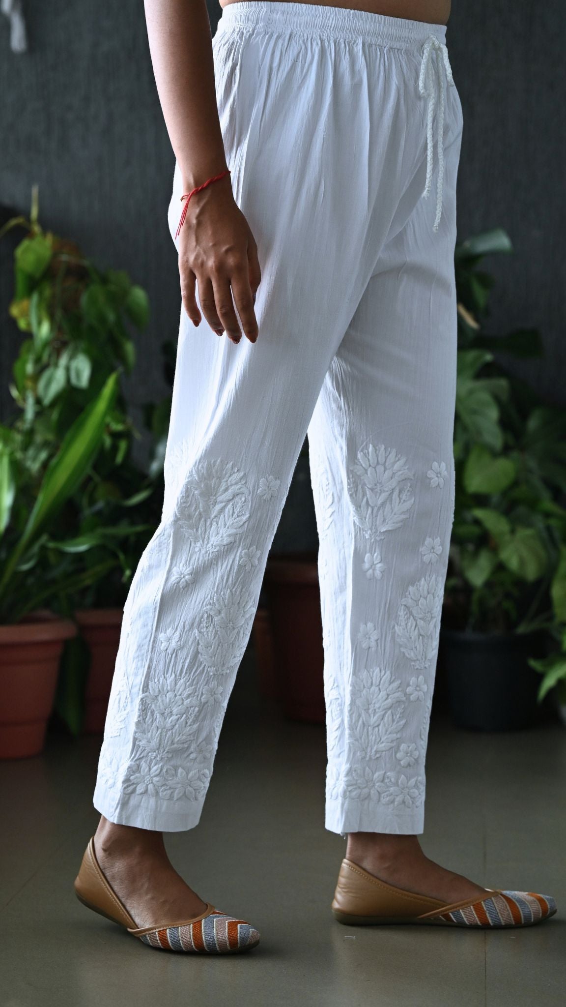 White embroidered cotton trousers with floral detailing on the lower legs, shown on a model wearing brown flat shoes in front of a dark wall with potted plants.