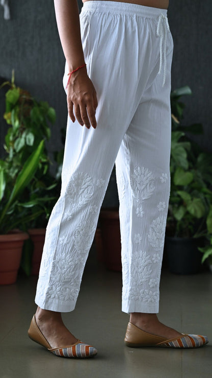 White embroidered cotton trousers with floral detailing on the lower legs, shown on a model wearing brown flat shoes in front of a dark wall with potted plants.