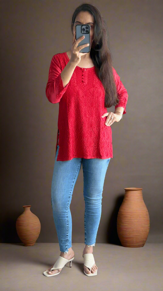 Person wearing a red chikankari top and blue jeans standing in front of a mirror with plants around.