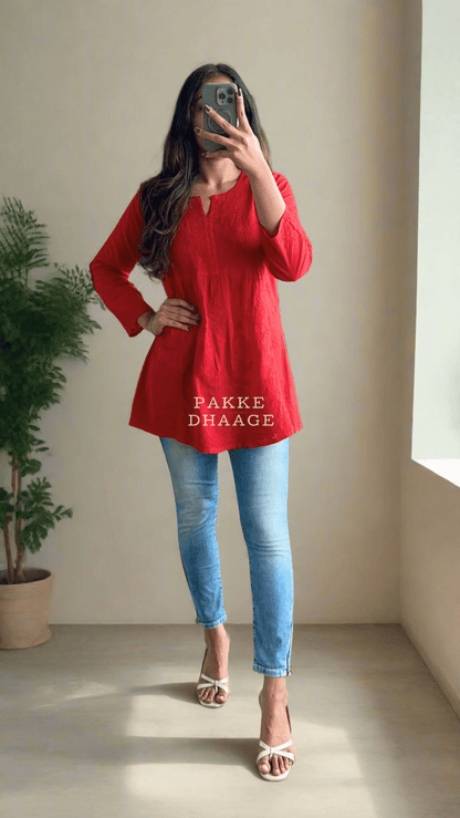 Person wearing a red top and blue jeans taking a mirror selfie in a plant-filled room.
