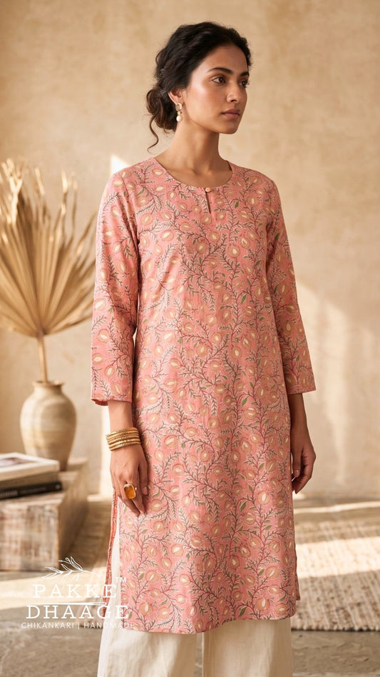 A woman wearing a dusty pink printed kurta with three-quarter sleeves and cream pants, standing in a warm, rustic indoor setting with a textured beige wall and dried palm leaves. | Pakke Dhaage
