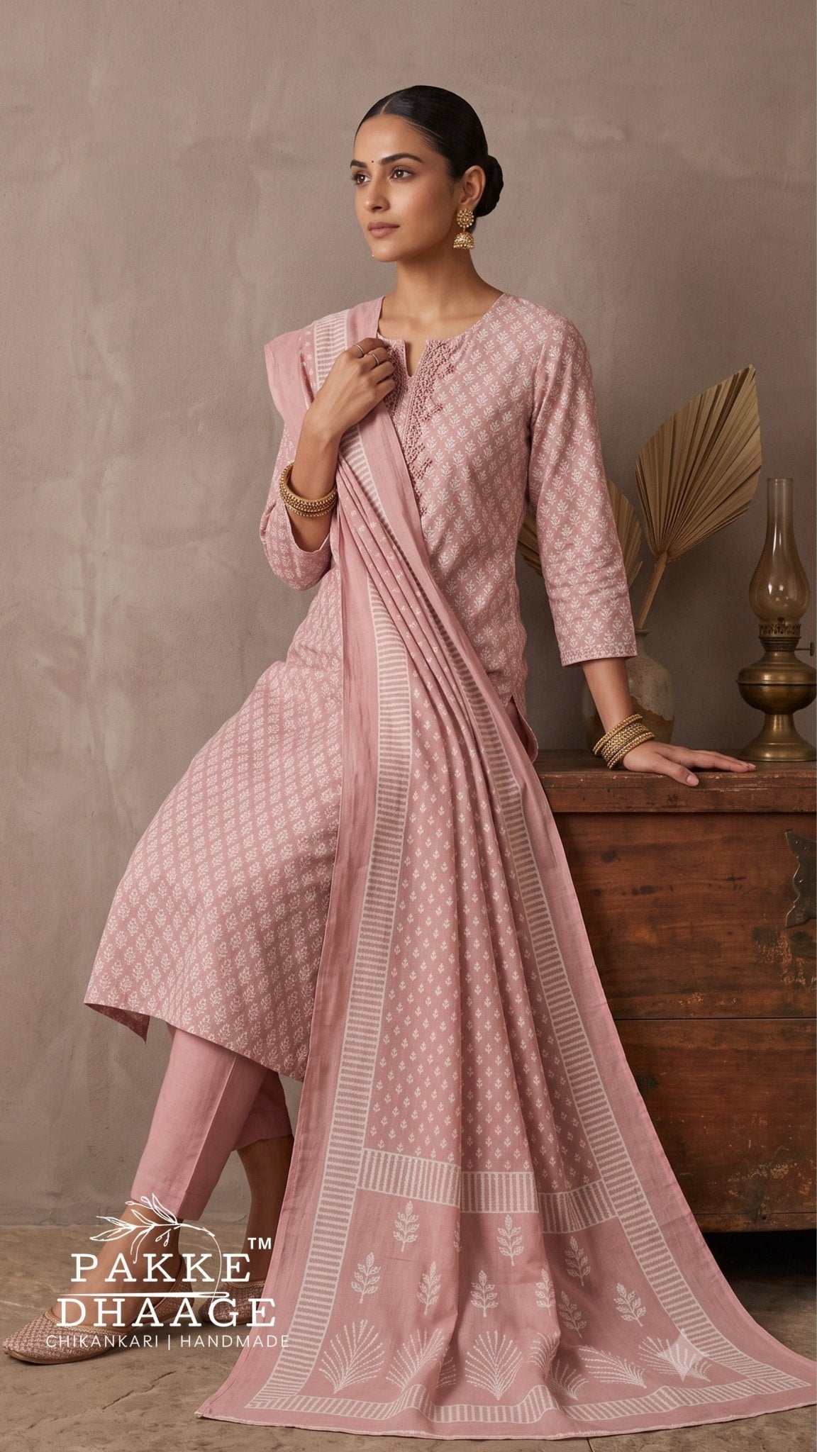 A woman models a light pink three-piece Anarkali suit set with a matching dupatta. The set features a subtle white geometric pattern, a solid pink inner pant, and a printed border on the dupatta.