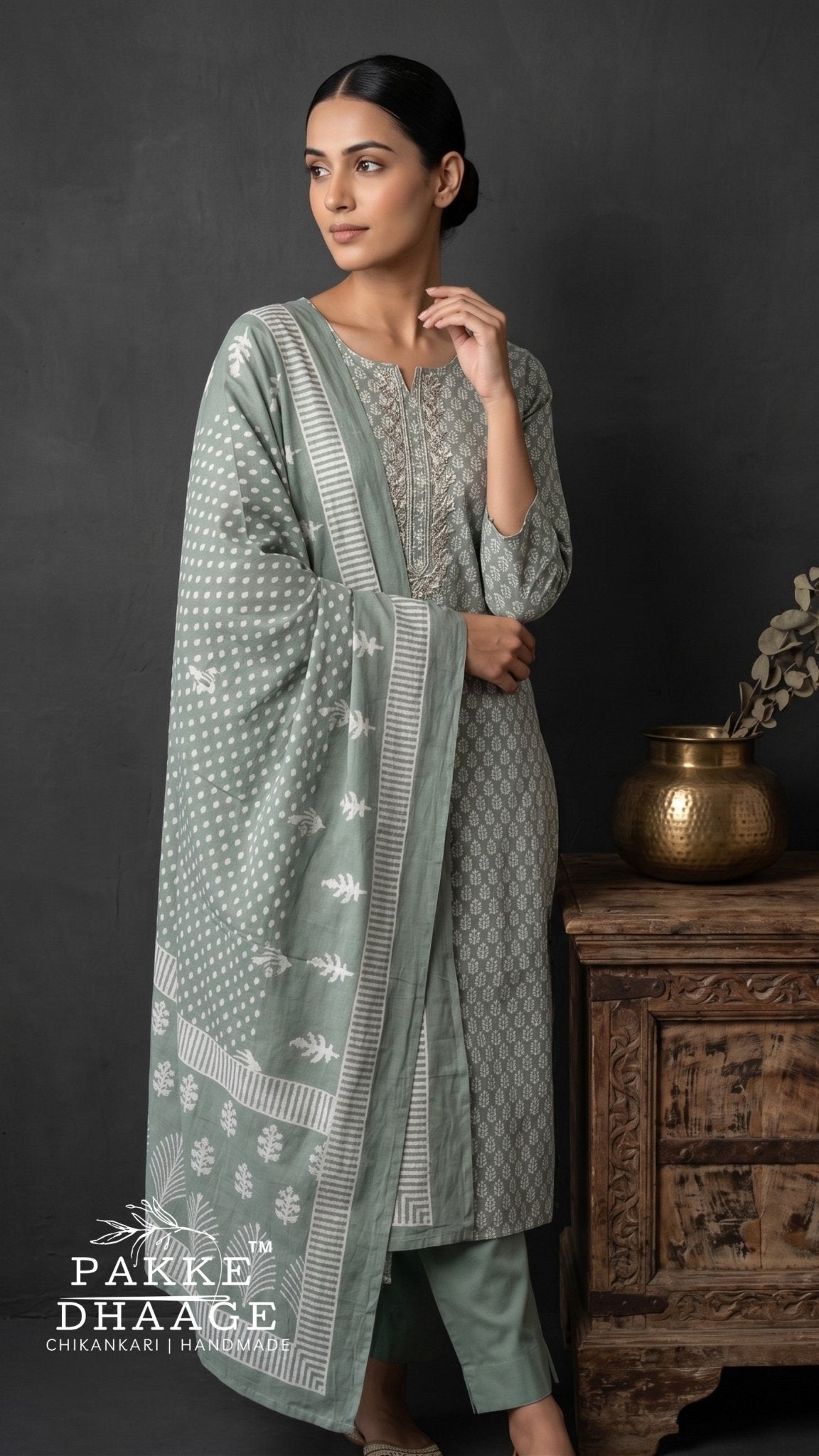 A woman models a three-piece traditional outfit in sage green and gray with white block prints. The set includes a long kurta, matching trousers, and a draped dupatta. | Pakke Dhaage