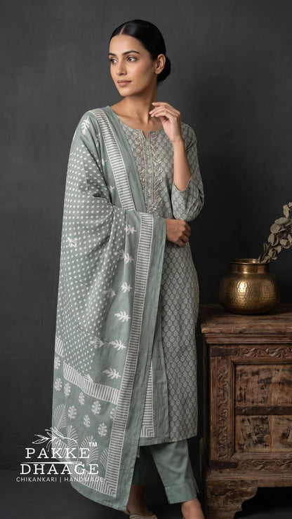 A woman models a three-piece traditional outfit in sage green and gray with white block prints. The set includes a long kurta, matching trousers, and a draped dupatta. | Pakke Dhaage