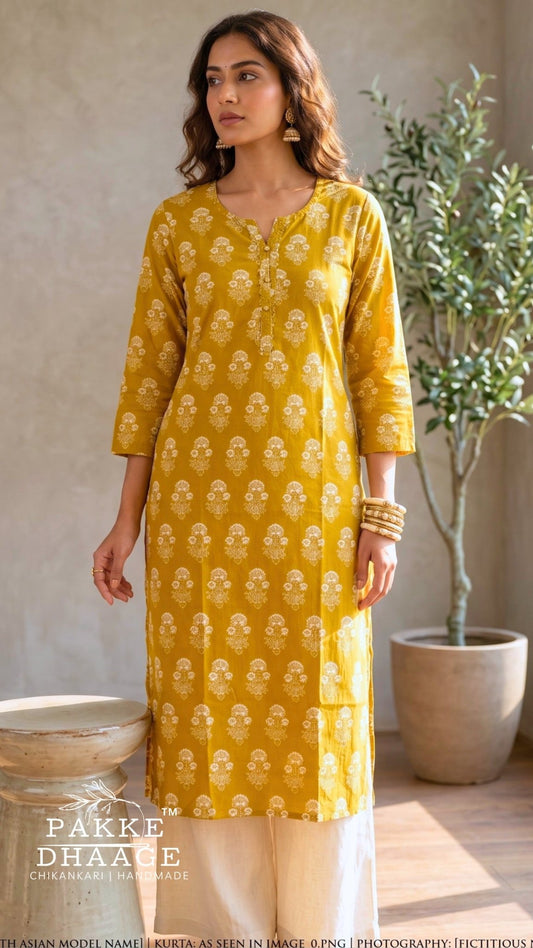 A woman modeling a mustard yellow kurta with white block prints, paired with cream trousers, in a styled indoor setting with a potted plant and rustic bowls. | Pakke Dhaage