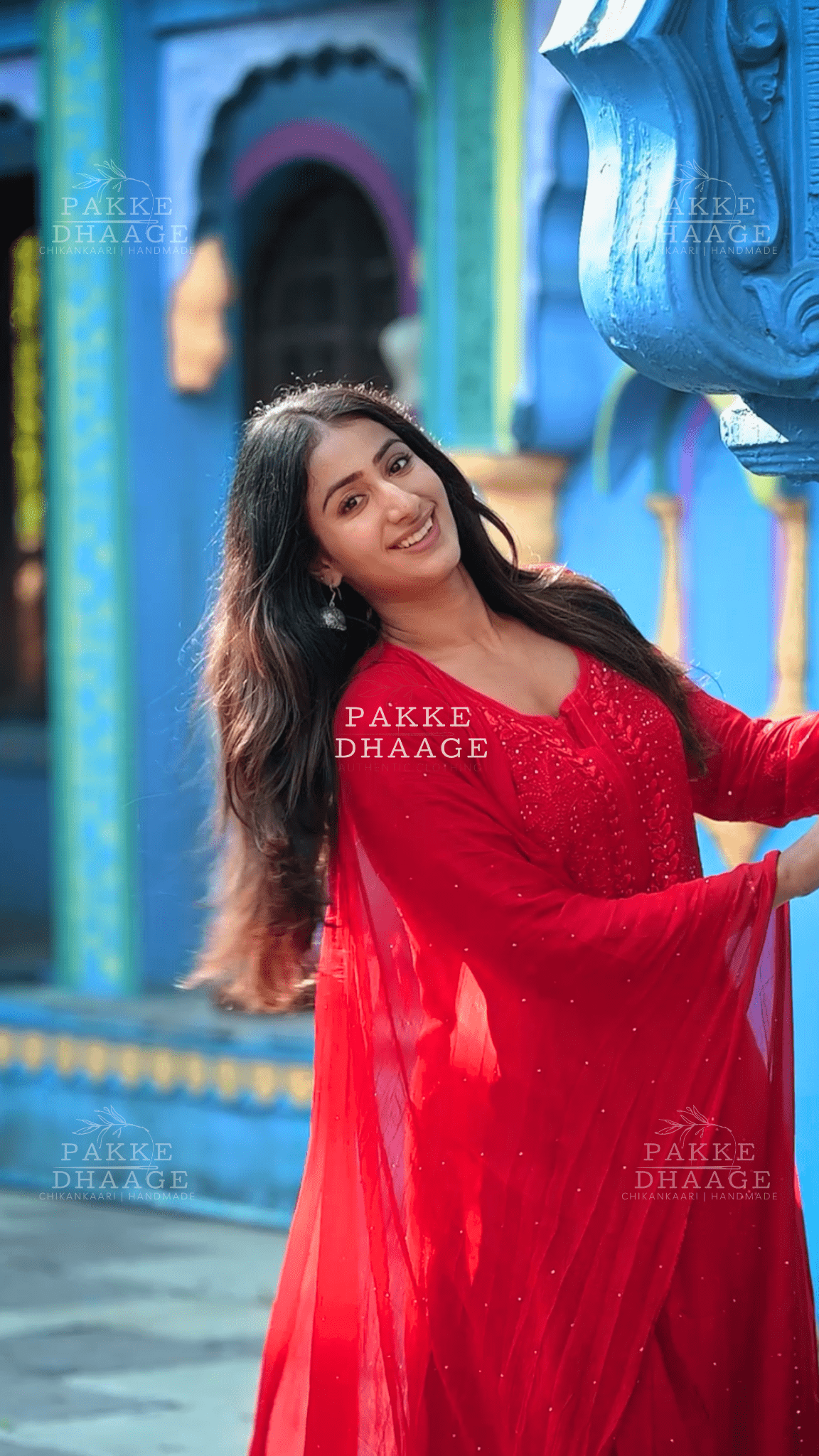 Woman in a red chikankari suit set, with handmade embroidery standing in front of a colorful architectural background with 'Pakke Dhaage' branding.