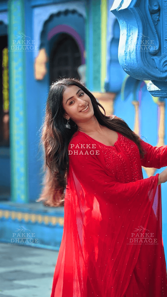 Woman in a red chikankari suit set, with handmade embroidery standing in front of a colorful architectural background with 'Pakke Dhaage' branding.