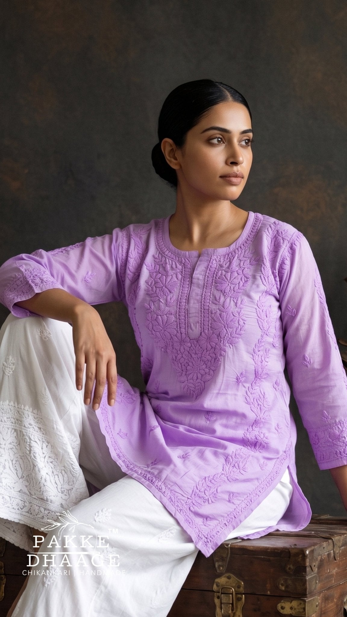 A woman models a lavender purple chikankari kurta with white pants, sitting on a rustic wooden trunk against a dark textured studio background. The brand name 'PAIKE DHARKE' is visible as a watermark in the bottom left corner.