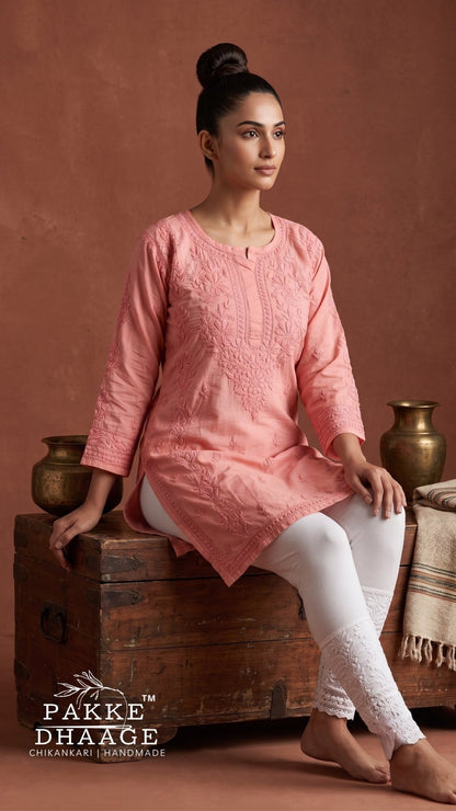 A woman seated on a rustic wooden crate wearing a peach-pink chikankari embroidered kurta with white pants, posed against a warm terracotta backdrop with decorative brass vessels. The brand name Pakke Dhaage Chikankari Handmade is visible in the lower left corner.