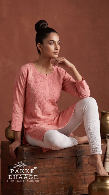 A woman models a pink long-sleeve chikankari kurta with white leggings while sitting on a rustic wooden chest against a warm terracotta background. The brand logo 'PAKE DHAAGE' is visible in the bottom left corner.