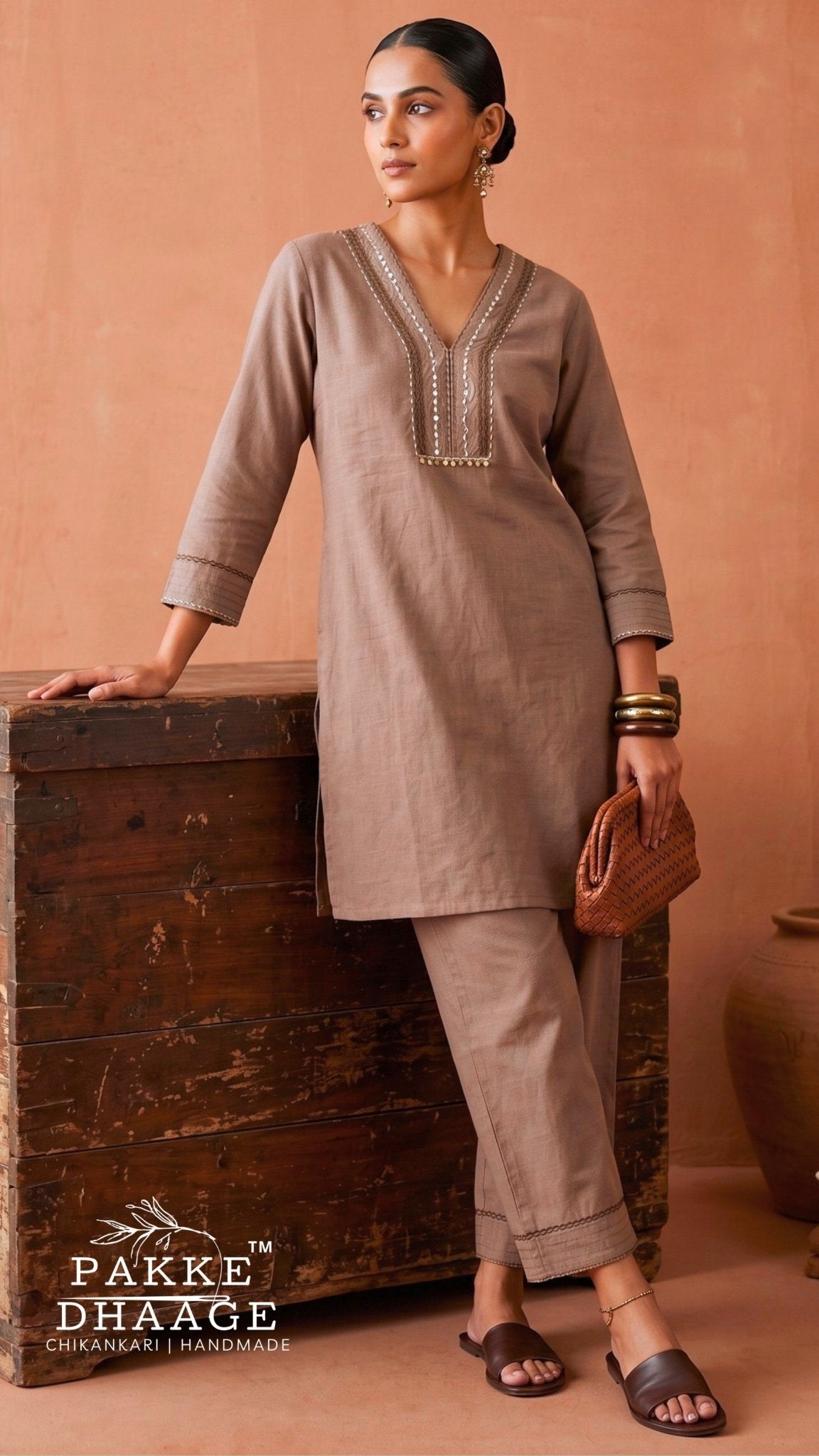 A woman models a taupe brown two-piece kurta and trouser set with silver embroidery on the neckline, standing next to a rustic wooden chest against a terracotta background. The brand logo 'PAkke DHAAGE' is visible in the bottom left corner.