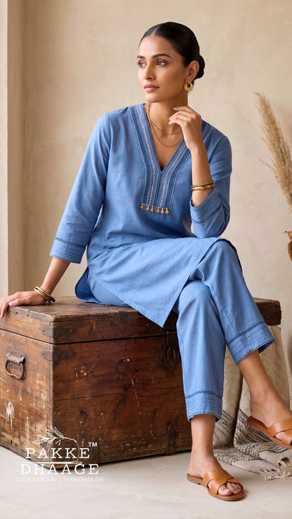 A woman wearing a light blue kurta and matching trousers sits on a rustic wooden chest in a warm, neutral-toned room. The brand name 'PAKE BHAGE' is visible as a watermark in the bottom-left corner.